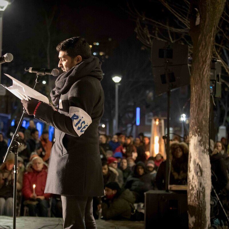 A person in a black coat and scarf speaks at a microphone at a candle lit vigil. They are wearing a white arm band that says "Rise."