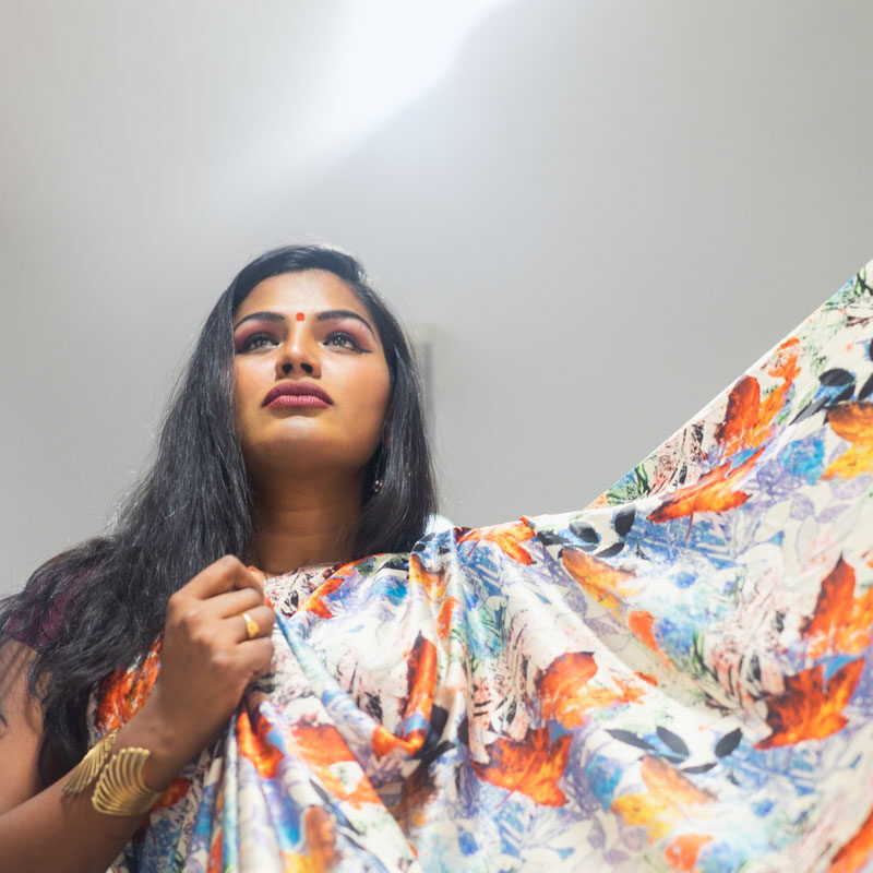 A trans woman with a bindi looks upward toward a glowing light source. She is holding a beautiful piece of colourful fabric and wearing gold jewelry.