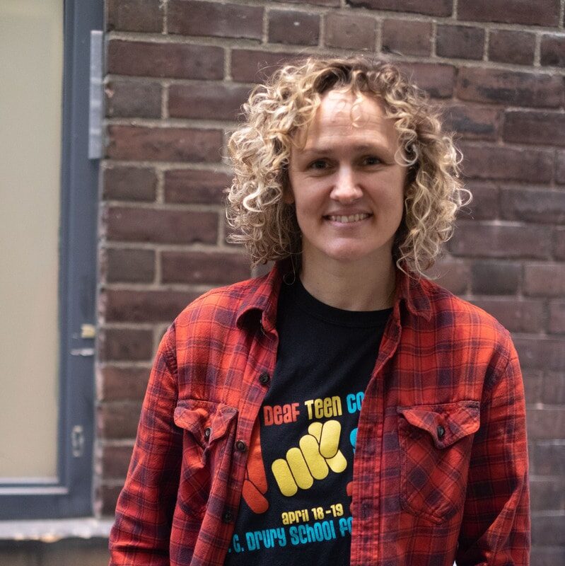 A person with blonde curly hair wearing a red plaid shirt smiles at the camera.