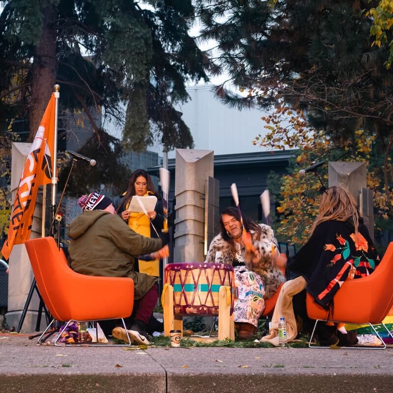 Three Indigenous drummers sit in a circle on orange chairs. They are all playing on a colourful drum and singing.