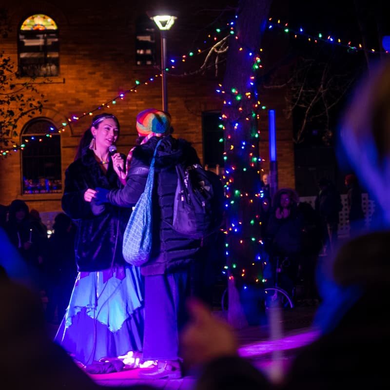 A person holding a microphone greets another person wearing a rainbow coloured hat. It's nighttime and they are standing beneath a tree covered in Christmas lights.