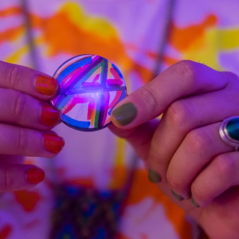 A close up of a person with colourful nails holding up a handmade button with a colourful "A" drawn in marker.
