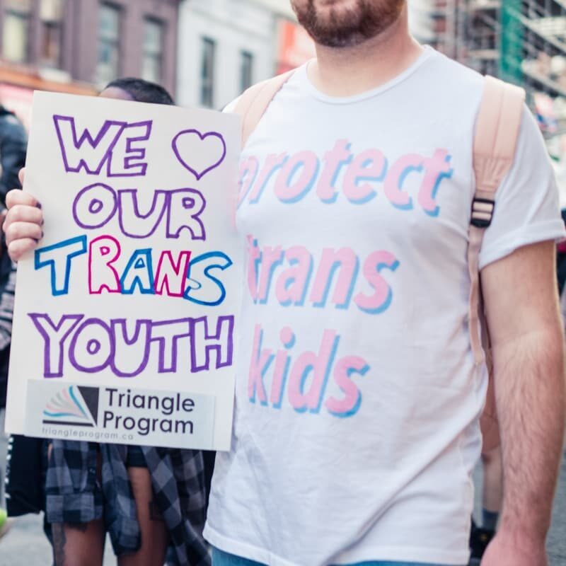 A person with a beard wearing a t-shirt that says "Protect Trans Kids" is holding a hand written sign that reads "We Love Our Trans Youth" followed by the Triangle Program logo.