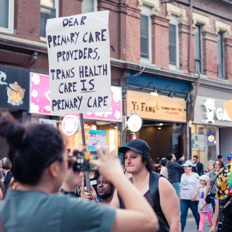 Peopling walking in Toronto's Trans March. A handmade sign that reads "Dear primary care providers, Trans health care IS primary care" is raised above the crowd.