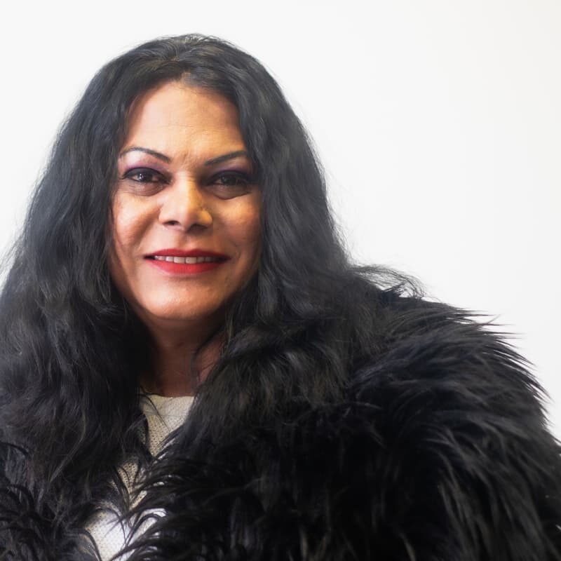 A member of The 519 Trans People of Colour project wearing a furry black coat and red lipstick is smiling at the camera.