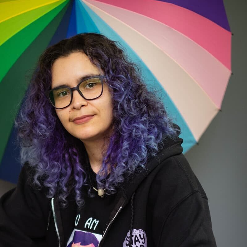 A 519 staff member smiling at the camera from beneath The 519's rainbow umbrella. They have curly purple hair and are wearing glasses and a black sweatshirt.