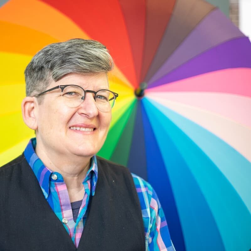 A 519 staff member with short grey hair and glasses smiling at the camera from beneath The 519's rainbow umbrella. They are wearing a plaid shirt in the colours of the Trans Flag and a black vest.