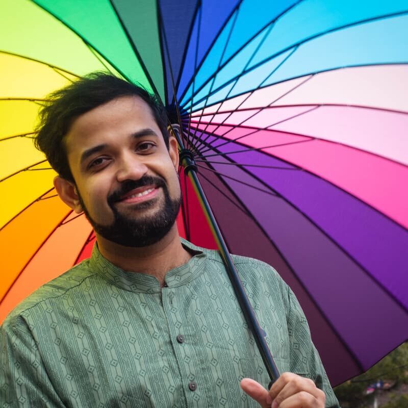 A 519 staff member with a dark beard and green shirt smiles at the camera from beneath The 519's rainbow umbrella.