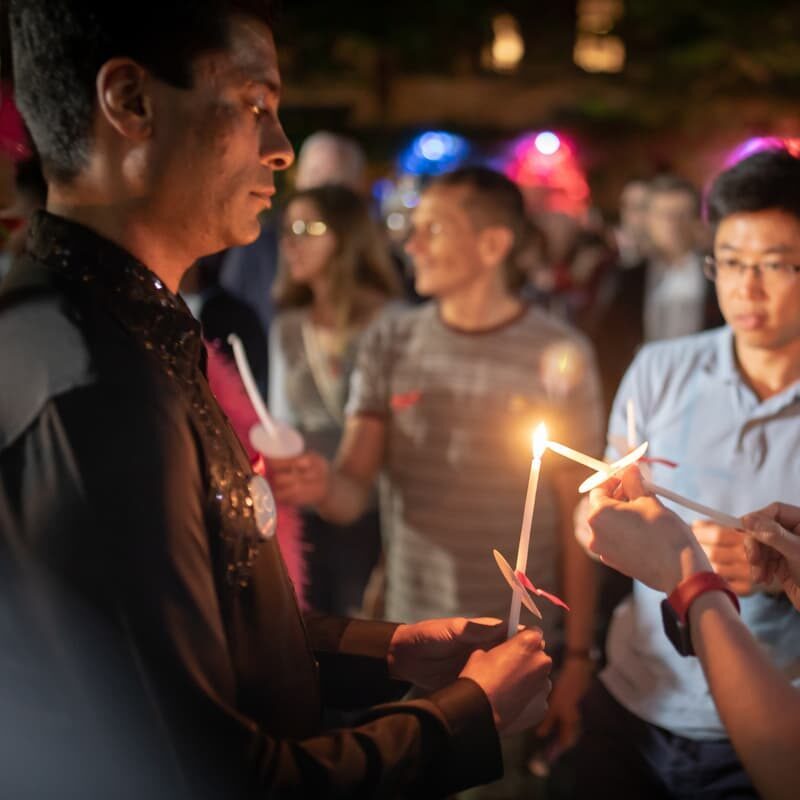 A group of people are attending a vigil. They are all holding candles tied with pink ribbons, and two people are using one candle to light another.