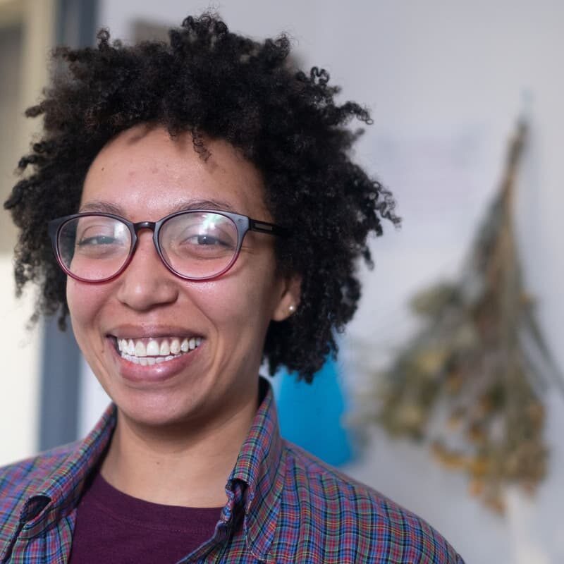 A 519 staff member with dark curly hair and glasses smiling at the camera. They are wearing a multi-coloured plaid shirt.