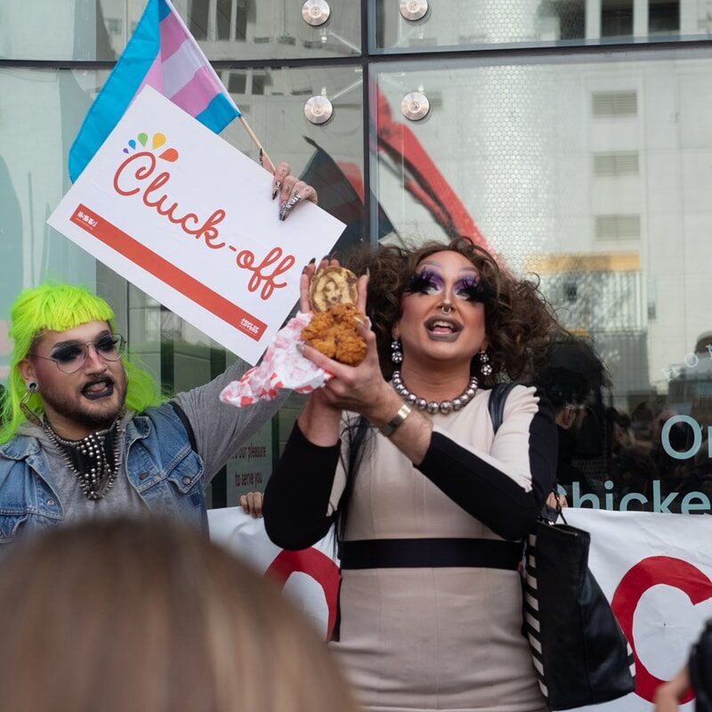 Two people in drag waving a trans flag and holding a sign that read "Cluck off" outside a Chick-fil-et in protest.