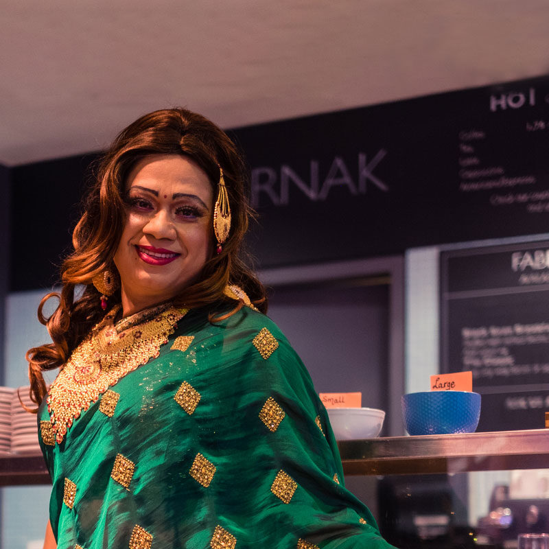 A member of The 519 Trans People of Colour project standing in The 519 kitchens. They are wearing a beautiful green sari, gold jewelry and a red bindi, and are smiling at the camera.